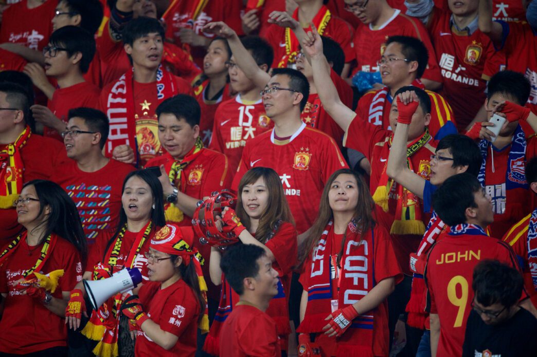 A large crowd of Chinese football fans, dressed in red and yellow, cheering loudly in a stadium.