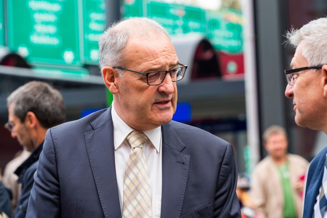Close-up of Guy Parmelin, wearing glasses and a suit, engaged in a conversation outdoors.