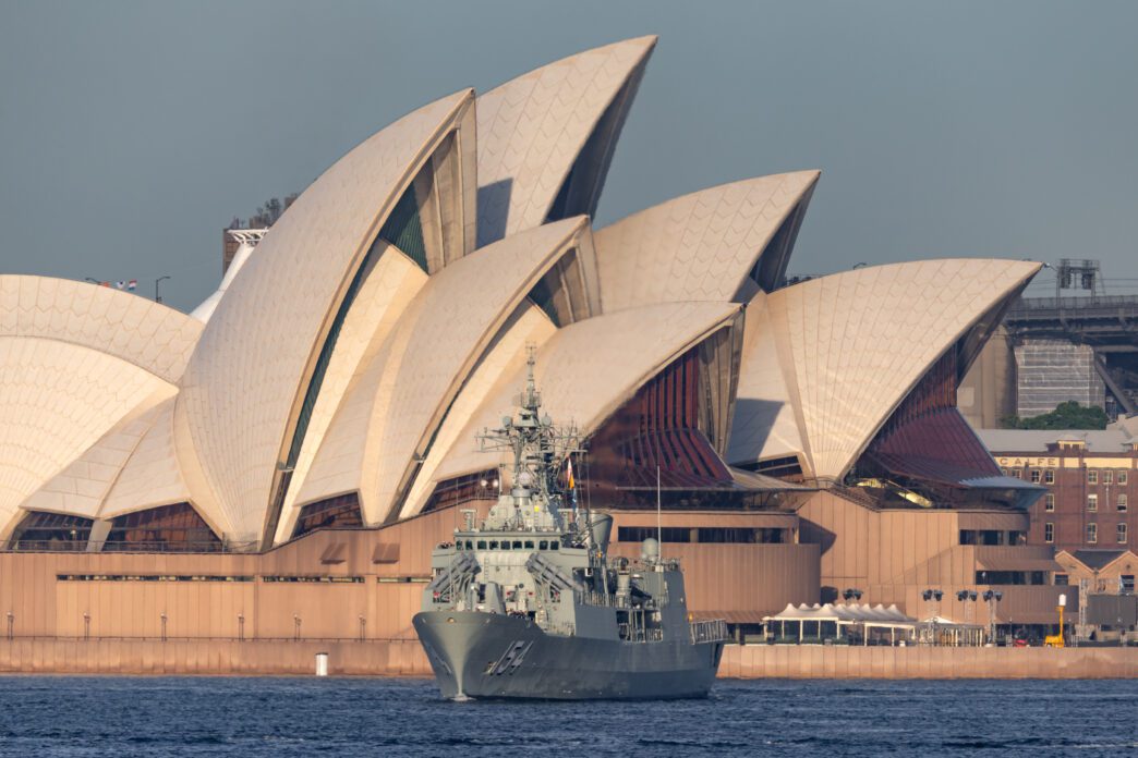The HMAS Parramatta frigate sails in Sydney Harbour with the iconic shell-like roofs of the Sydney Opera House visible in the background.