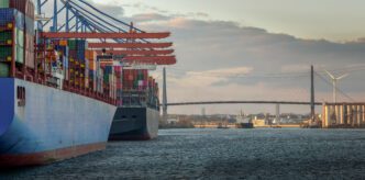 Two large container ships docked in the Port of Hamburg with the Köhlbrand Bridge in the background at sunset.