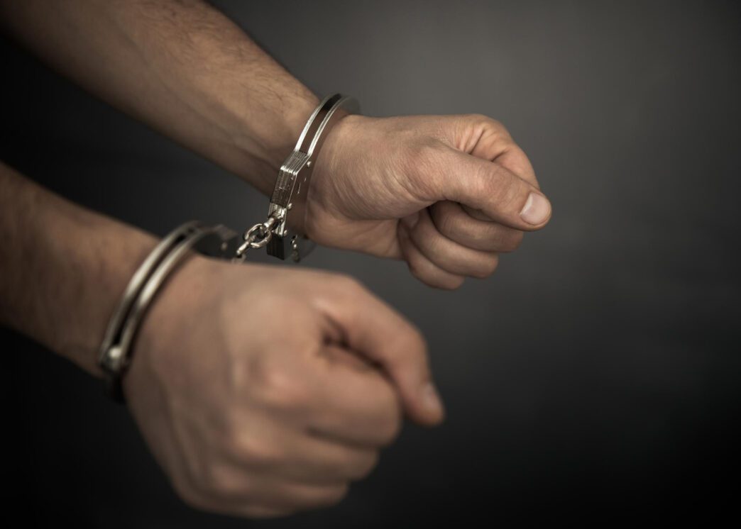 Close-up of a person's hands tightly bound together with shiny metal handcuffs against a dark background.