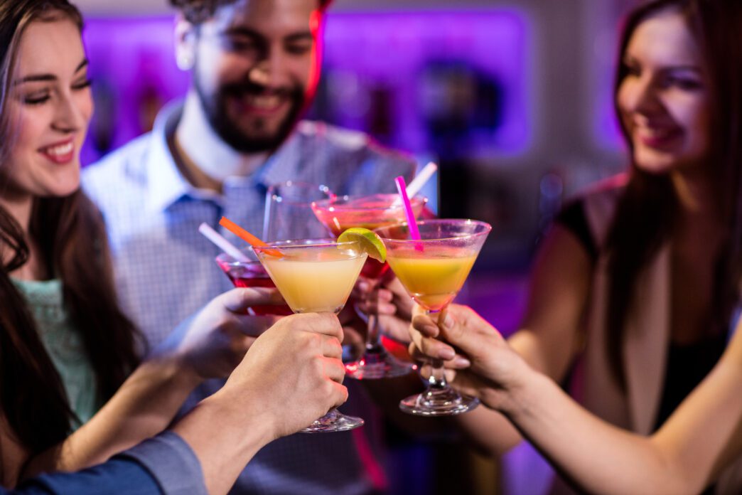 Group of friends happily toasting with colorful cocktails at a brightly lit bar.