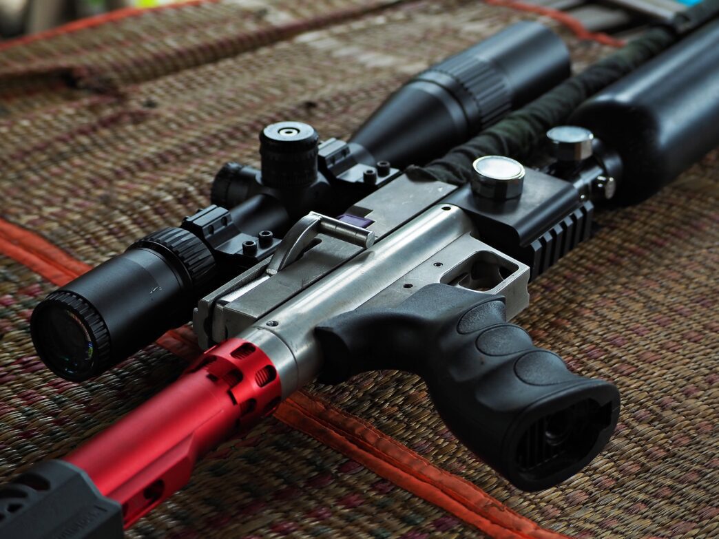 Close-up of a tactical air rifle with a scope and a red barrel on a woven mat.