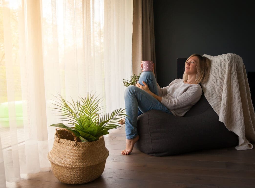 Woman relaxing barefoot in a beanbag chair near a sunny window and a large potted palm plant.