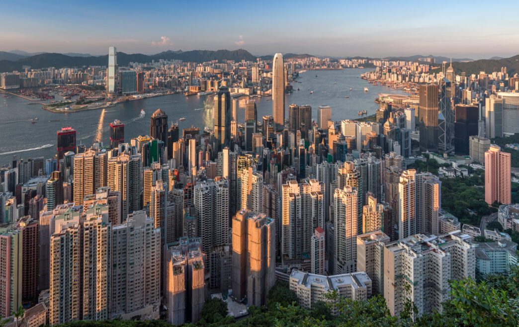 Sweeping aerial panorama of the densely packed Hong Kong skyline with skyscrapers, apartment buildings, and Victoria Harbour at sunset.