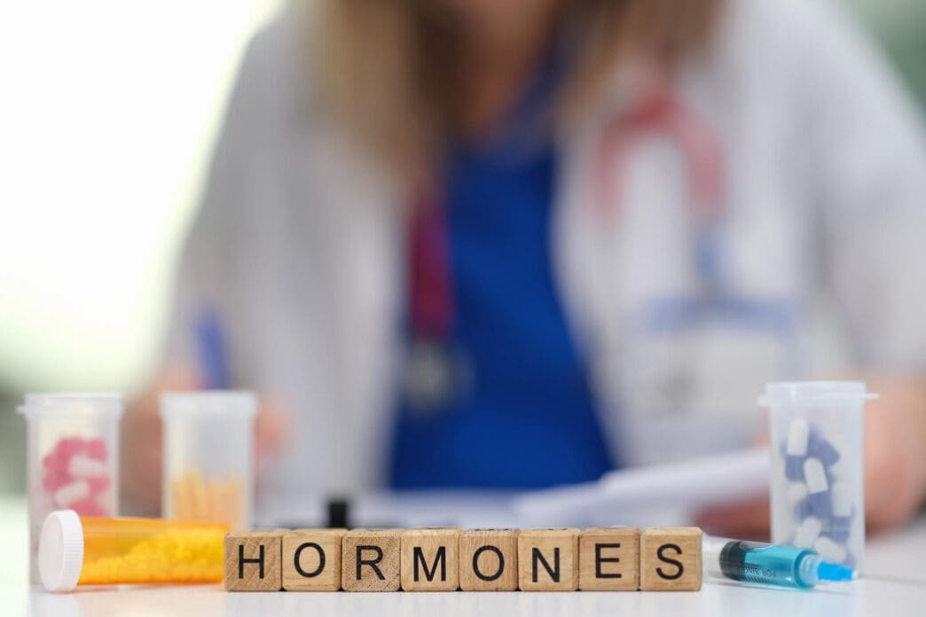 Wooden blocks spelling "HORMONES" sit in the foreground, with medication bottles and a blurry person in a lab coat behind them.