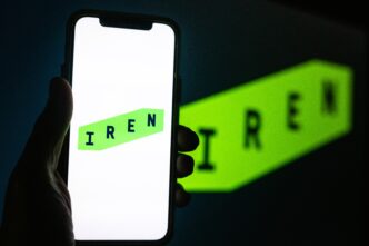 Hand holding a smartphone displaying the green and white IREN logo in front of a computer screen showing a larger, blurred version of the same logo.