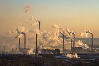 Industrial factory skyline with smoke billowing from multiple tall smokestacks at sunset.
