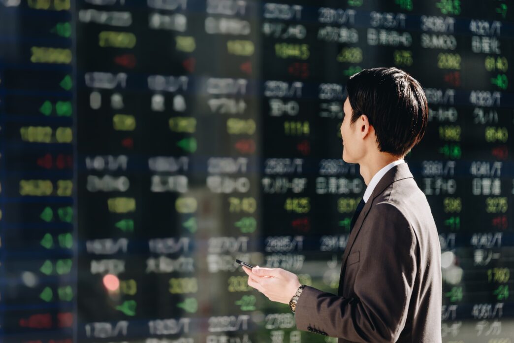 Japanese businessman in a suit checking stock prices on a large, reflective electronic ticker board while holding a smartphone.