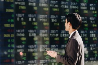 Japanese businessman in a suit checking stock prices on a large, reflective electronic ticker board while holding a smartphone.