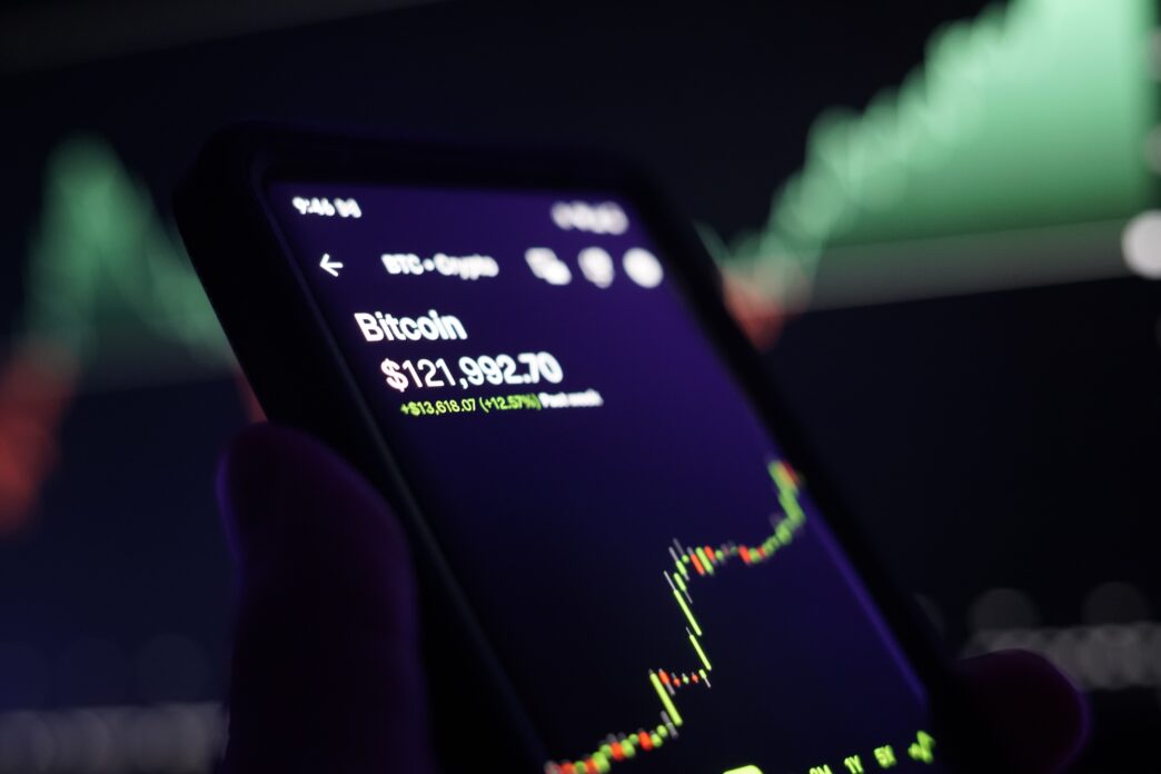 Smartphone screen displaying the Bitcoin price at $121,992.70 and a candlestick chart showing a sharp rise in value.
