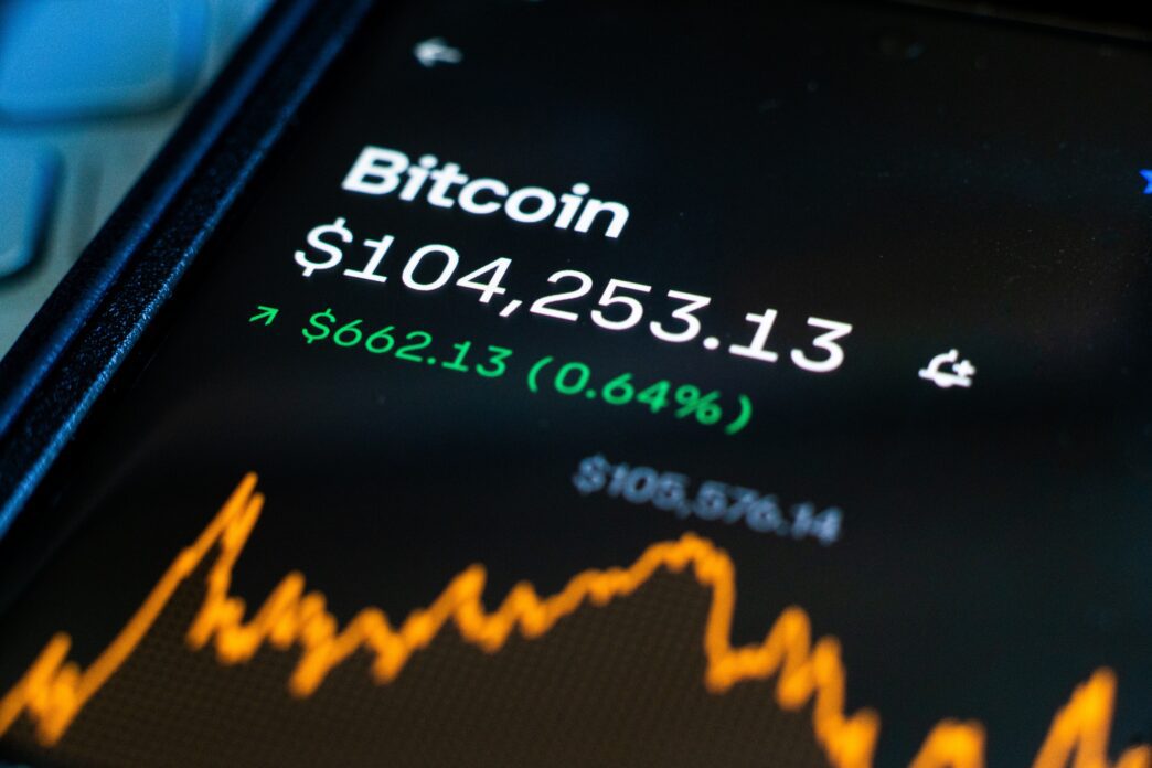 Close-up of a smartphone screen showing the live Bitcoin price at $104,253.13 and a price increase of 0.64% over an orange line chart.