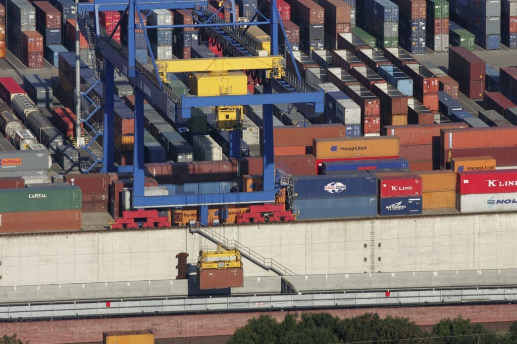 Aerial view of a large container terminal, showing a blue and yellow gantry crane moving containers.