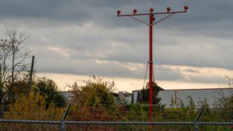 Red airport approach lighting tower and damaged industrial building under cloudy, evening skies.