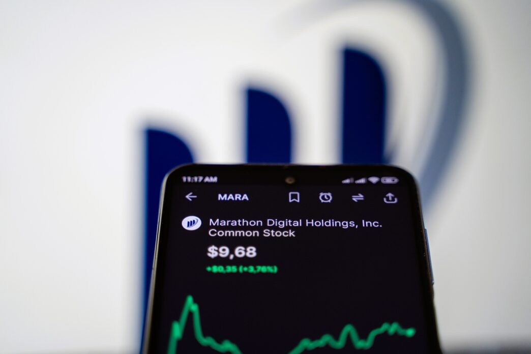 Smartphone screen displaying the stock price and graph for Marathon Digital Holdings (MARA) against a blurred corporate logo backdrop.