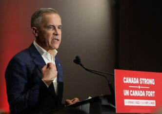 Mark Carney, in a suit, speaks forcefully at a podium with a red sign reading "CANADA STRONG."