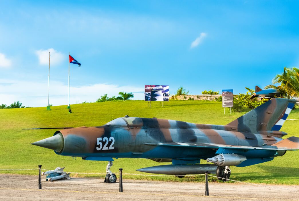 Camouflaged Cuban MiG fighter jet displayed outdoors on a grassy hill under a bright blue sky.
