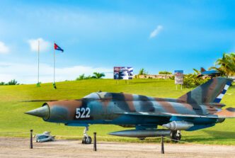Camouflaged Cuban MiG fighter jet displayed outdoors on a grassy hill under a bright blue sky.