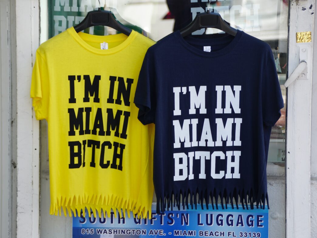 Two novelty T-shirts, one yellow and one navy, displaying the phrase "I'M IN MIAMI BITCH" on a store window.