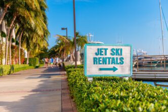 A turquoise and white sign for "Jet Ski Rentals" with an arrow pointing right, along a sunny, palm-lined Miami boardwalk.