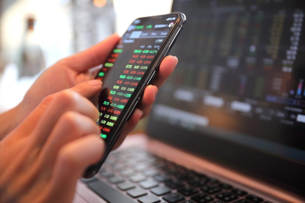 A person's hands holding a smartphone displaying stock market data, with a laptop in the background.