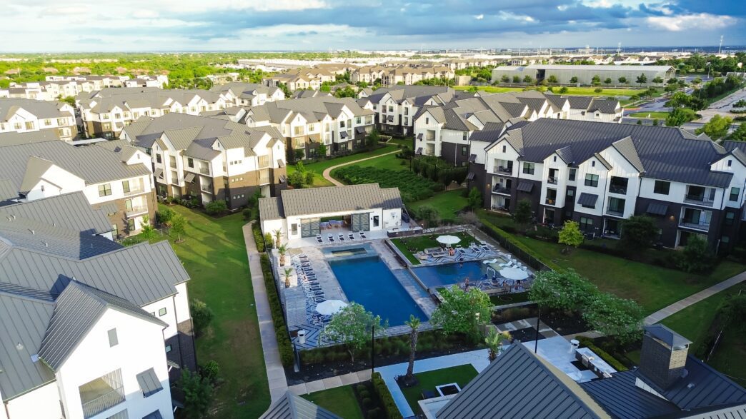 Aerial view of a modern apartment complex with metal roofs, surrounding a large swimming pool and patio area.