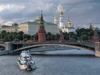 A river cruise boat passing under a bridge with the Spasskaya Tower and Moscow Kremlin in the background.