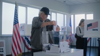 Elderly man drops ballot into a transparent box in a polling station with US flags.