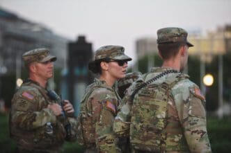 Three members of the US Army National Guard in camouflage uniform and tactical gear stand outdoors at dusk.