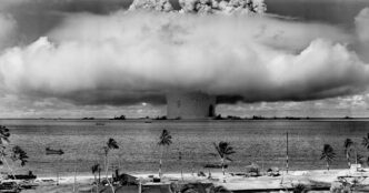 Black and white panoramic photograph of a massive nuclear mushroom cloud rising from an ocean surface near a palm-tree-lined beach.
