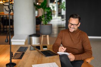 Smiling man in glasses and a brown sweater taking notes while working on a laptop in a modern office.
