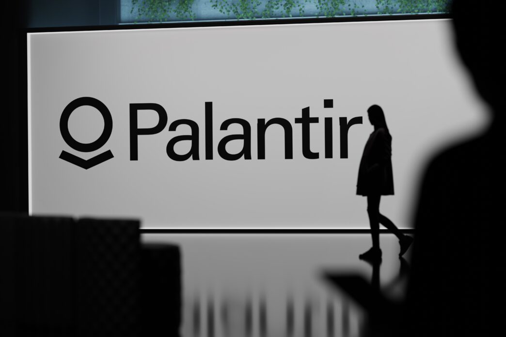 Silhouette of a person walking past a large screen displaying the Palantir Technologies logo.