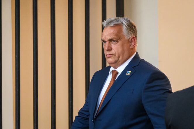 Close-up of Hungarian Prime Minister Viktor Orban, wearing a blue suit and orange tie, with a serious expression.