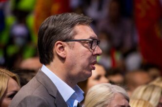 Profile close-up of Serbian President Aleksandar Vucic wearing glasses and speaking at a political campaign rally.
