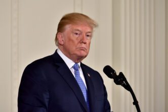 President Donald Trump stands at a microphone in a dark suit and blue tie, delivering remarks at a White House press conference.