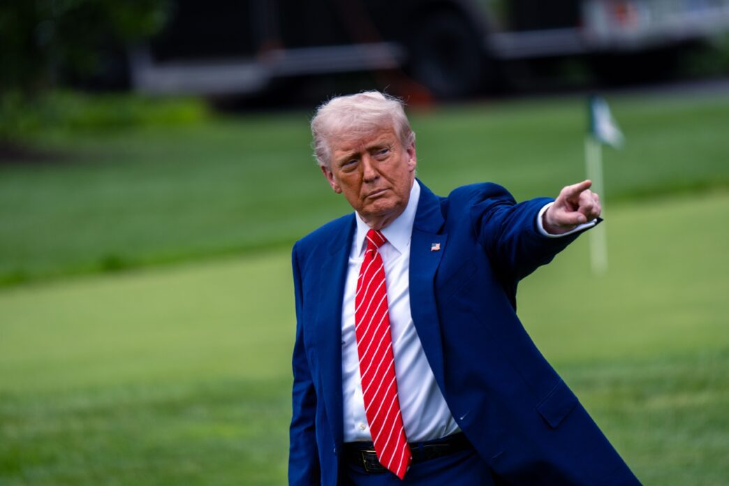 President Donald Trump, in a blue suit and red tie, strides across a green lawn and points to the right.