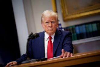 President Trump in a blue suit speaking at a microphone in the White House Roosevelt Room.