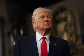 President Donald Trump in a dark suit and red tie, looking slightly upward in a formal indoor setting.