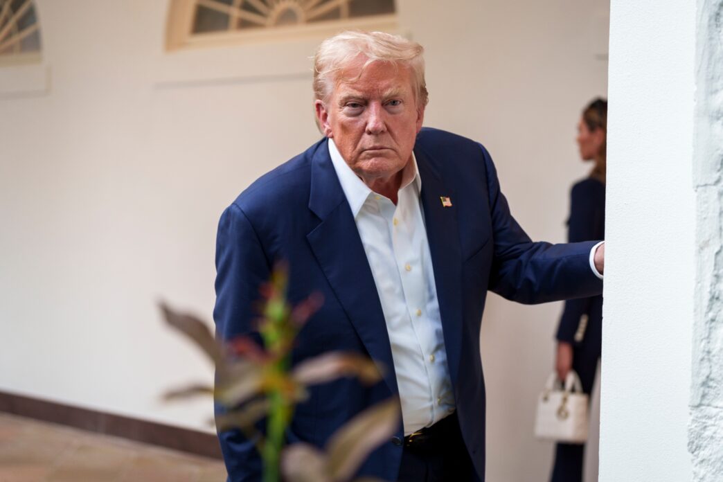 President Trump, in a blue suit, standing near a White House doorway with a serious expression.