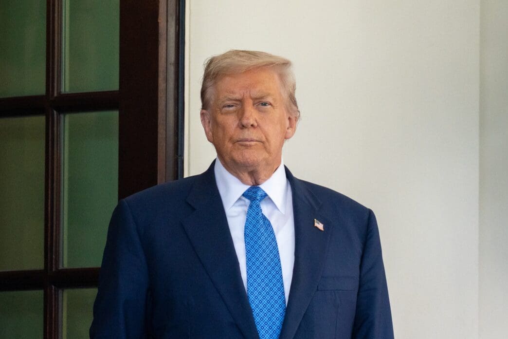 President Donald Trump stands formally in a navy suit and blue tie, looking directly at the camera.