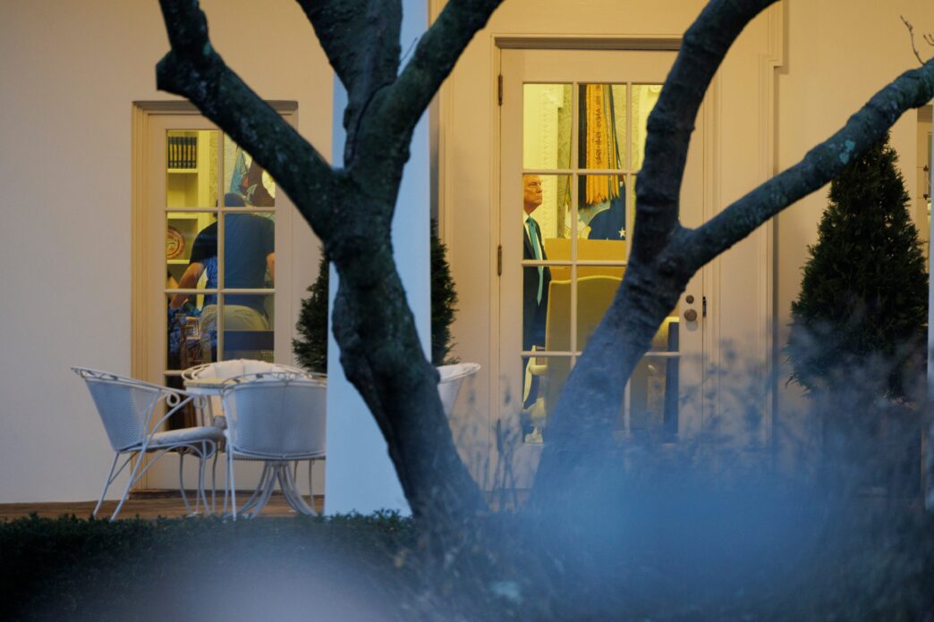 President Trump seen through the glass door of the brightly lit Oval Office at dusk.