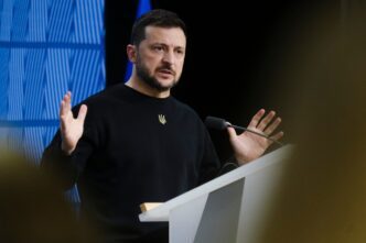 Ukrainian President Volodymyr Zelenskiy addresses a press conference, gesturing with both hands while wearing a dark sweater with a trident emblem.