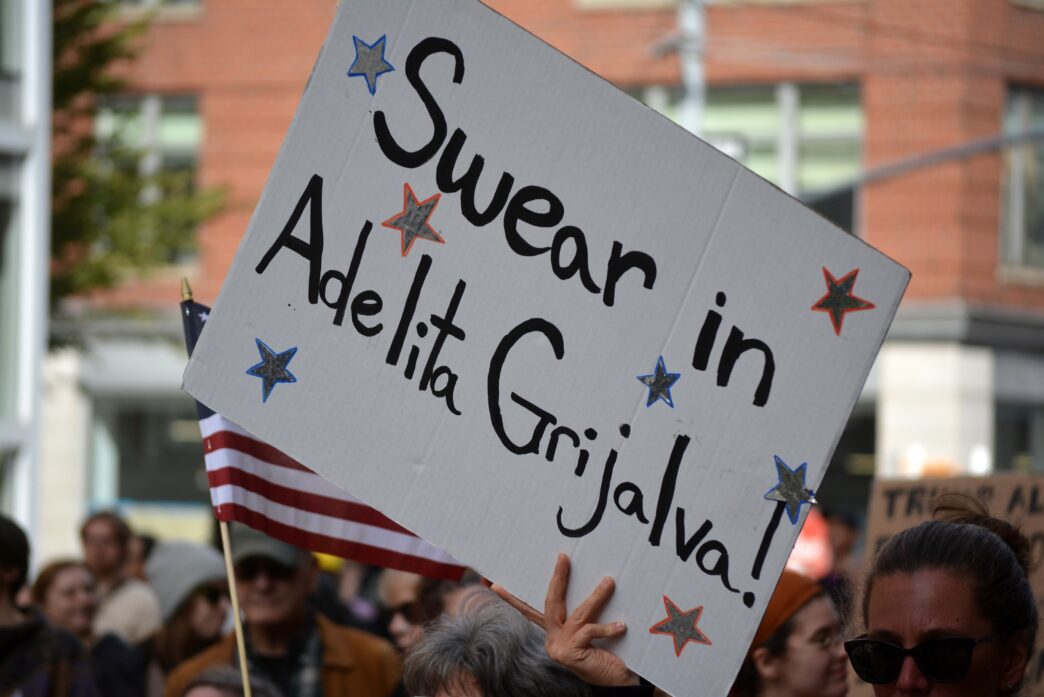 A protest sign reading "Swear in Adelita Grijalva!" with a small American flag visible below it.