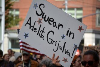A protest sign reading "Swear in Adelita Grijalva!" with a small American flag visible below it.