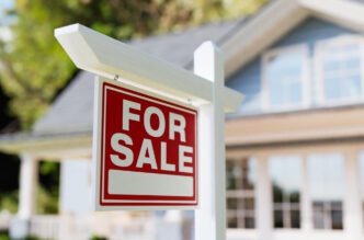 A red and white "For Sale" sign on a wooden post in front of a blurred, traditional suburban house.