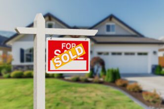 Real estate sign with a red "For Sale" portion covered by a yellow "SOLD" sticker in front of a house.