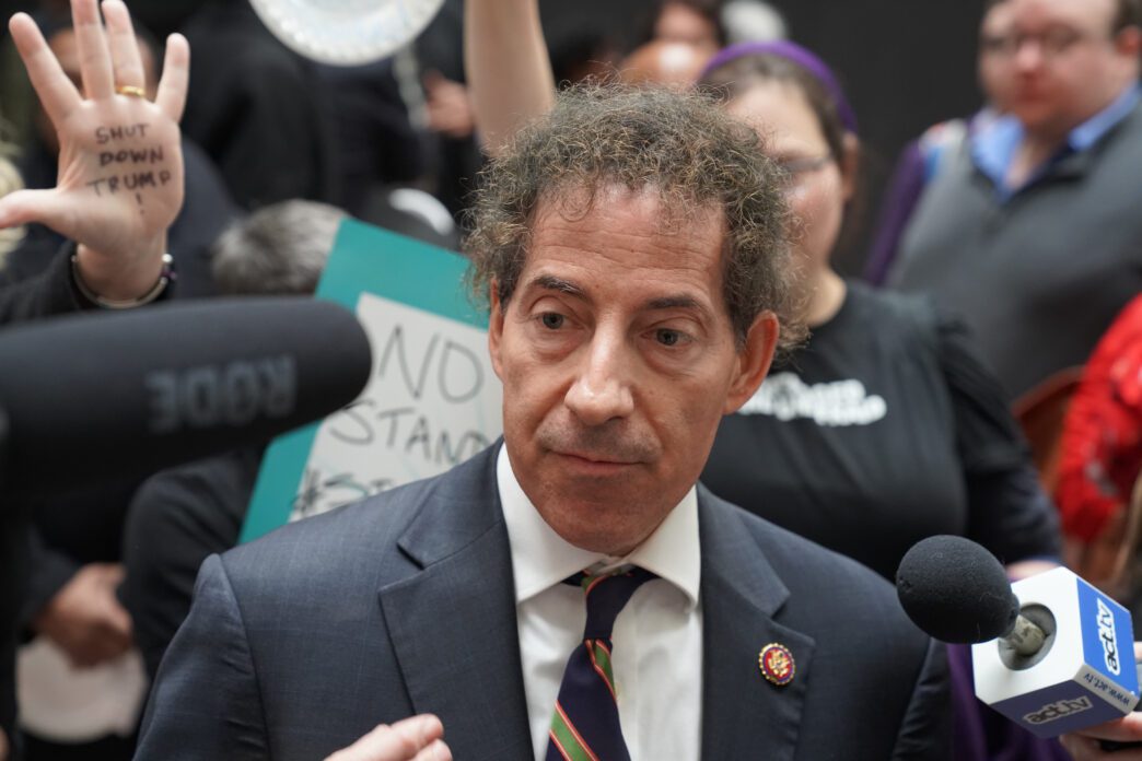 Rep. Jamie Raskin speaking to reporters, surrounded by protesters holding signs and an anti-Trump message.