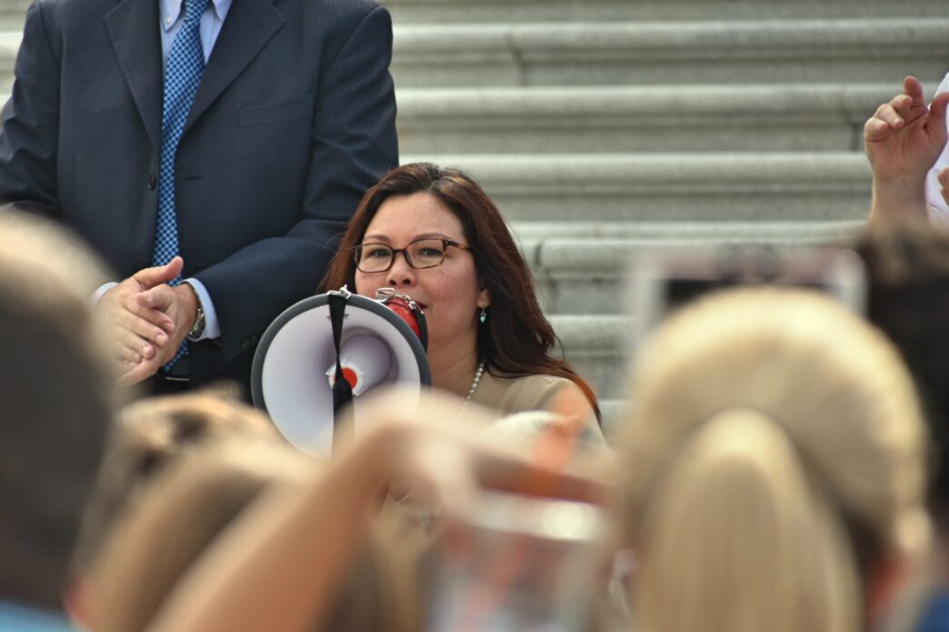 Senator Tammy Duckworth speaking into a megaphone to supporters on the steps of the US Capitol.