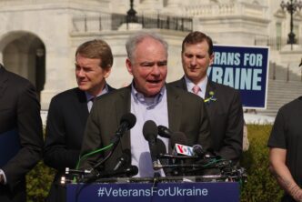 U.S. Senator Tim Kaine speaking at a press conference outdoors, with a "Veterans for Ukraine" sign in the background.