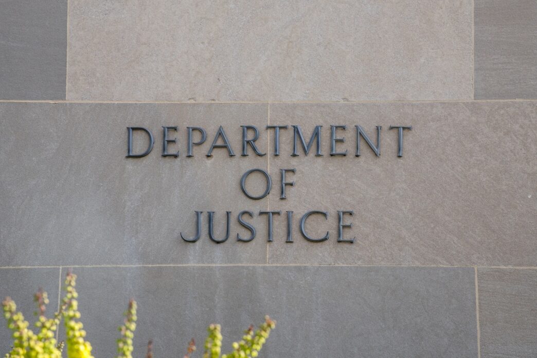 Close-up of raised, dark metallic letters reading "DEPARTMENT OF JUSTICE" on a beige stone wall.